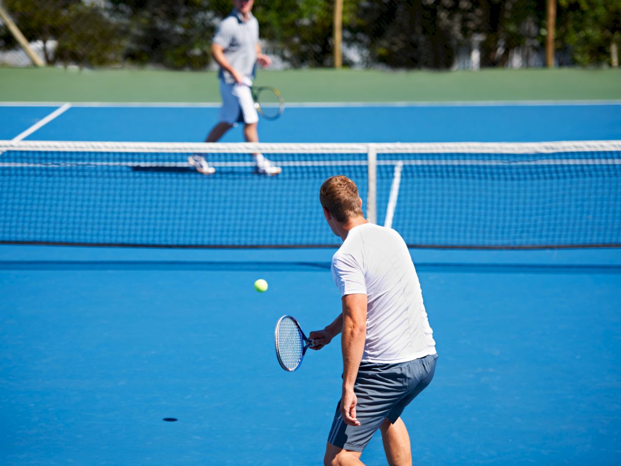 The image features two men playing tennis on a blue court, with one preparing to hit the ball while the other positions himself.