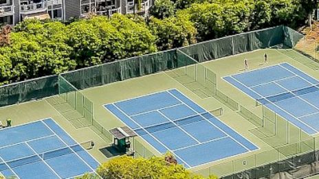 The image shows multiple tennis courts with blue surfaces, surrounded by greenery and a few players visible on the courts.