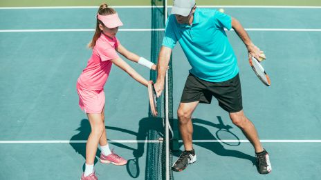 A girl in a pink outfit and a man in a turquoise shirt play tennis, touching rackets across the net on a court.