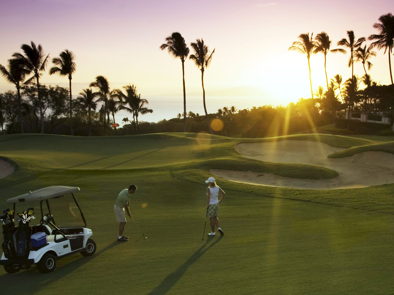 The image shows two golfers on a lush course at sunset, with palm trees and a golf cart in view. A serene, picturesque scene.