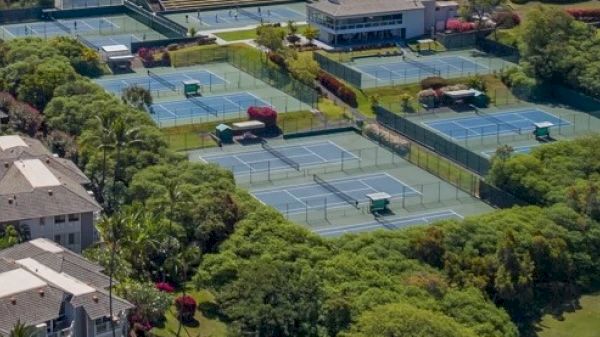 The image shows an aerial view of multiple tennis courts surrounded by greenery and a building nearby. It look like a recreational area.