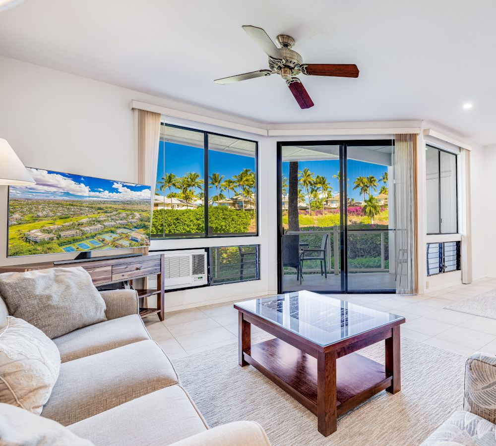 A bright living room with a ceiling fan, TV, sofa, coffee table, and large windows showcasing a view of palm trees and greenery outside.