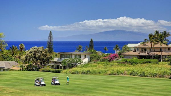 The image shows a scenic golf course with two golf carts, a player, ocean views, and lush green surroundings under a blue sky.