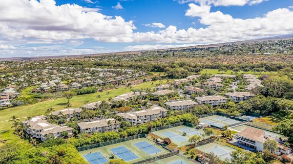 The image shows an aerial view of a residential area with green spaces and several tennis courts below a blue sky.