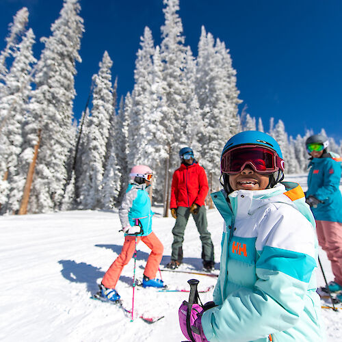 The image shows a group of people skiing in a snowy landscape, surrounded by tall trees under a clear blue sky. They're having fun.