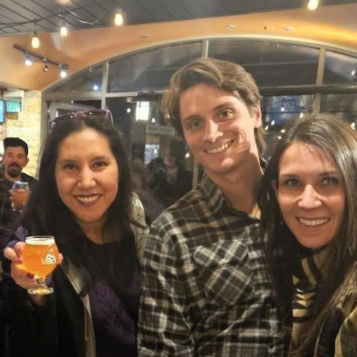 The image shows three smiling people standing together, with one holding a drink in a lively indoor setting.