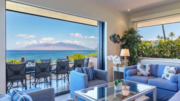 The image shows a stylish living room with ocean views, featuring blue couches, a glass table, and sliding doors to an outdoor area.