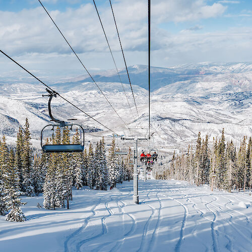 A snowy mountain landscape with ski lift chairs, tall pine trees, and distant hills under a clear blue sky invites winter adventures.