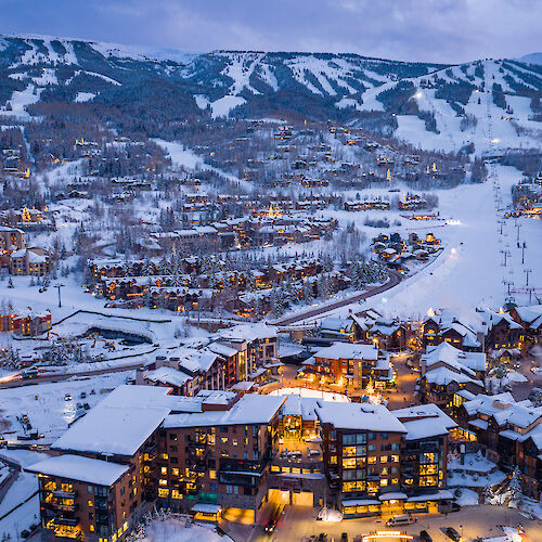 The image shows a snowy mountain landscape with ski slopes, a cozy resort, and illuminated buildings at dusk. It's scenic and inviting.
