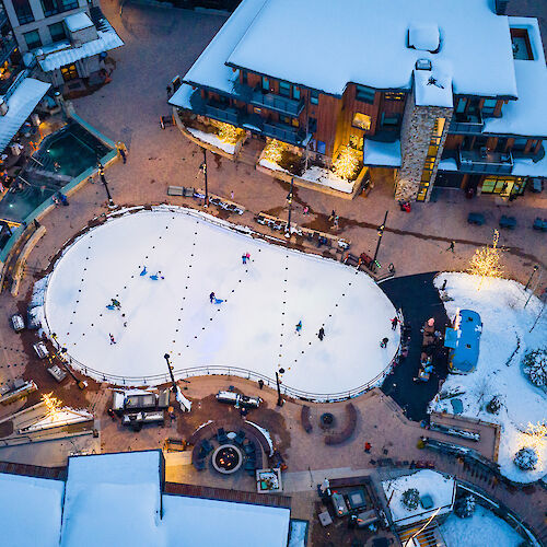 The image shows a snowy resort area with a skating rink and buildings around it, bustling with activity in a winter setting.