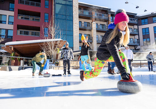 A girl curls on ice in colorful pants while others enjoy the sport in a sunny, modern outdoor setting. Winter fun abounds.
