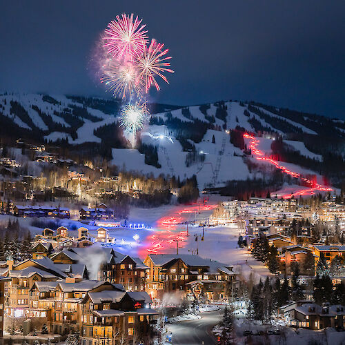 The image shows a night scene in a snowy village with colorful fireworks in the sky and illuminated buildings. It looks festive.
