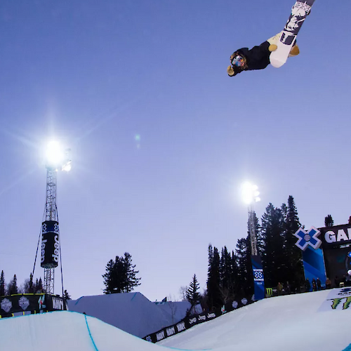A snowboarder performs an aerial trick at a nighttime event, illuminated by bright lights, with spectators in the background.