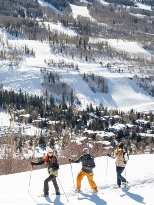 Three people skiing on a snowy slope with a mountainous landscape and a town visible in the background. It&rsquo;s a sunny day.