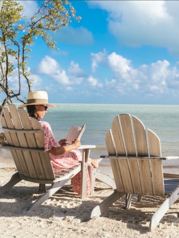 A woman reads on a beachside, seated in an Adirondack chair, with a serene ocean view and a clear blue sky above.