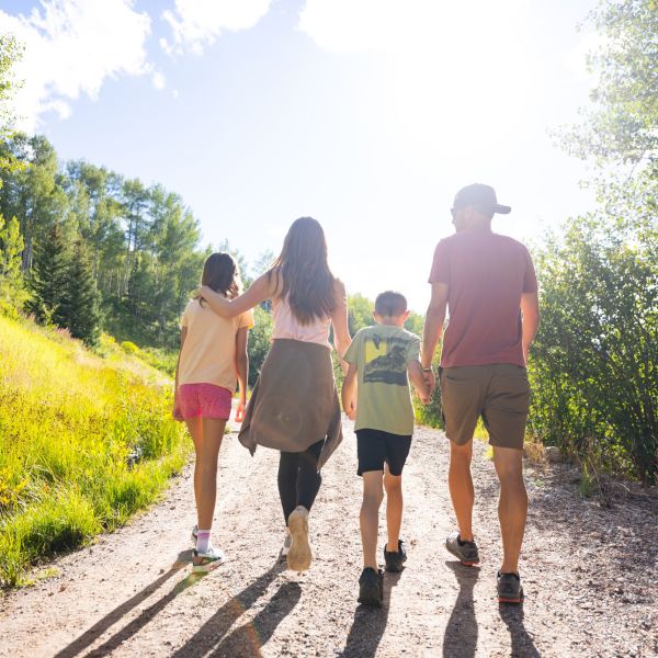 A family hikes up a sunlit trail together through a green, sunny landscape, from behind as they walk.