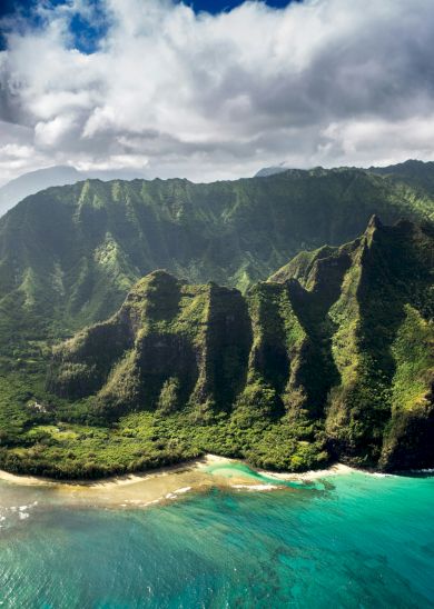 Aerial view of lush green cliffs meeting turquoise ocean, with a rocky coastline and clear blue sky above, showcasing tropical coastal scenery.