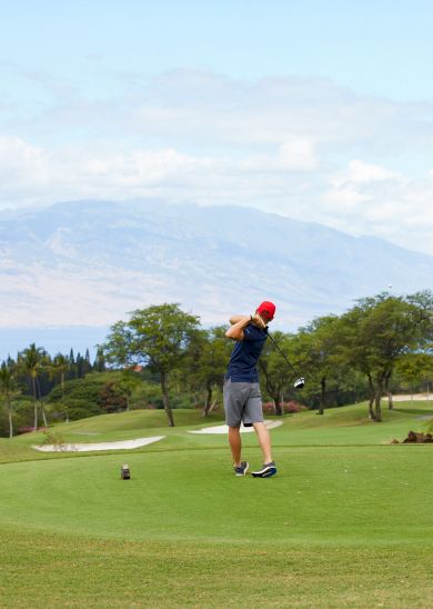 A golfer is swinging on a course with mountains in the background, surrounded by greenery and open fairways.