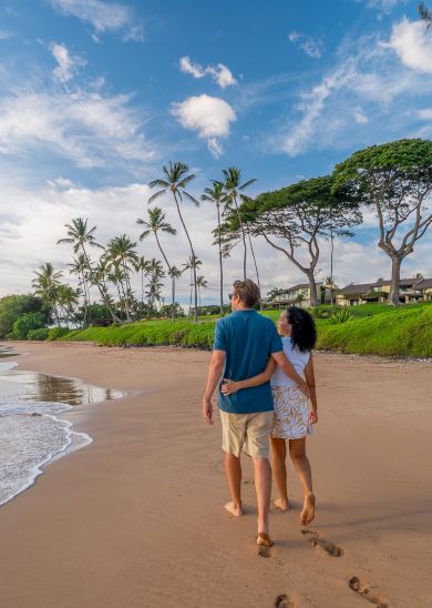 A couple walking hand-in-hand along a sandy beach with palm trees, gentle waves, and a bright, partly cloudy sky.
