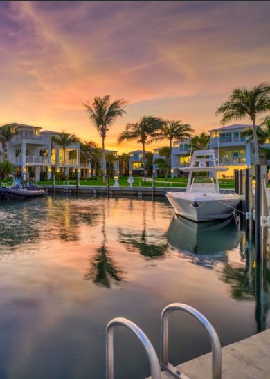 The image depicts a serene waterfront scene at sunset with palm trees, homes, and a boat reflecting on calm waters.