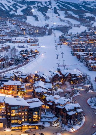 The image shows a snowy mountain village with ski slopes, cozy buildings, and winding roads, all illuminated by evening lights.
