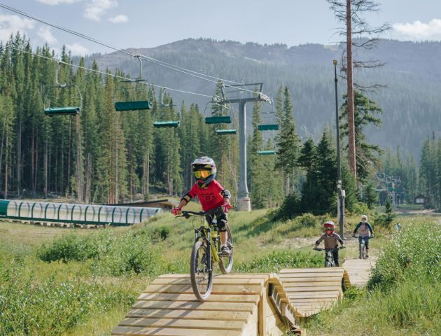 Three people are mountain biking on a trail with ramps, surrounded by trees and mountains. A lift system can be seen in the background.