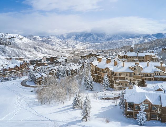 A picturesque, snowy mountain village with wooden buildings, snow-covered trees, and winding roads, set against a backdrop of mountains and a blue sky.