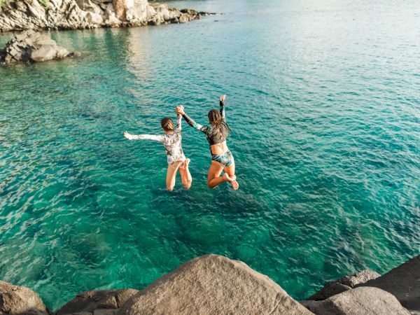 Two people are mid-jump into clear blue water from a rocky shore, holding hands and wearing swimwear, with a sunlit coastline in the background.