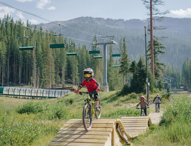 A young cyclist rides over wooden ramps in a scenic forested area, with ski lifts and friends in the background.