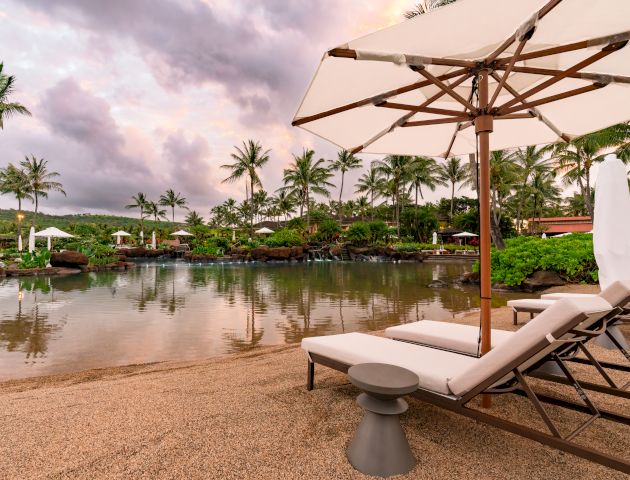 A serene beach scene featuring lounge chairs and an umbrella by a calm lagoon surrounded by palm trees and a cloudy sky.