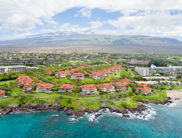 The image depicts a coastal area with buildings and lush greenery, featuring ocean waves and a backdrop of mountains under a cloudy sky.