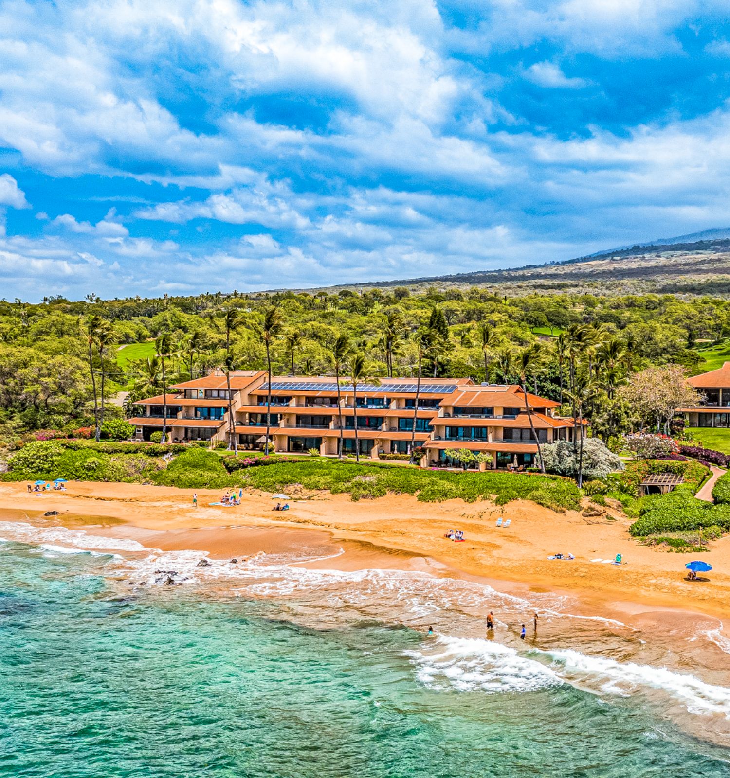 The image shows a scenic beach with clear water, sandy shore, and lush greenery, featuring large buildings along the coastline.
