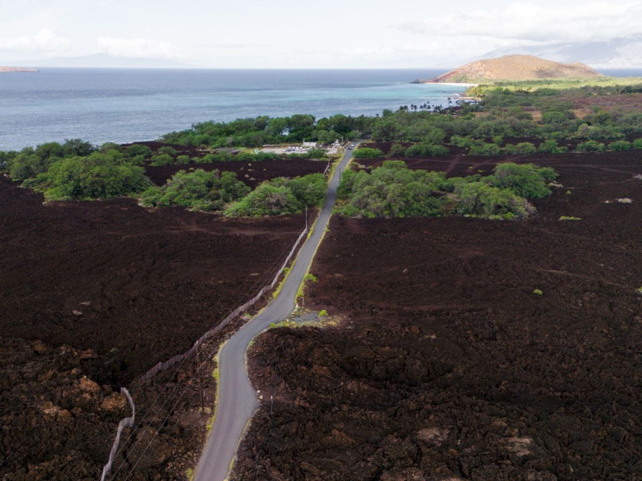 The image shows a coastal area with a scenic road surrounded by black lava fields and green vegetation, near the ocean.