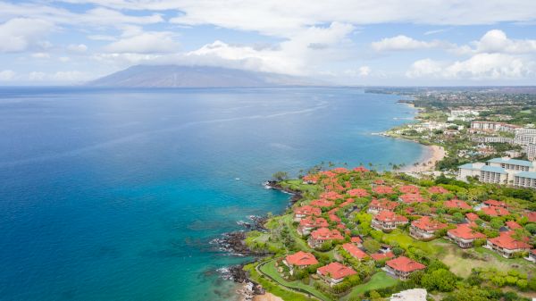 The image shows a scenic coastal view with lush greenery, red-roofed buildings, and clear blue waters, likely in a tropical setting.