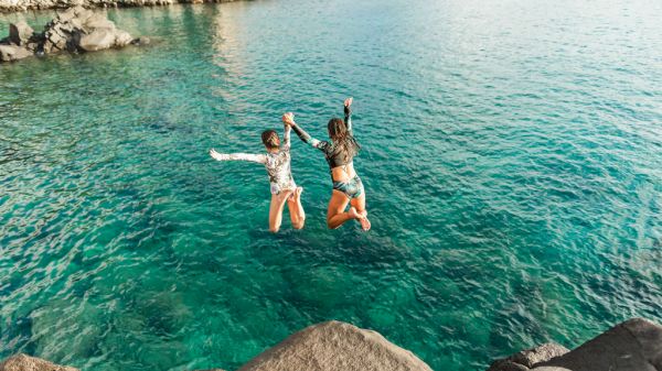 Two people are joyfully jumping into clear blue water from a rock, capturing a moment of excitement and fun.