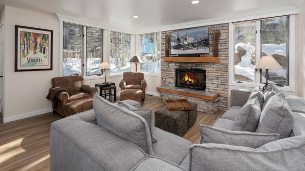 A cozy living room with a gray couch, armchairs, a stone fireplace, and large windows showing a snowy outdoor scene. Artwork decorates the wall.