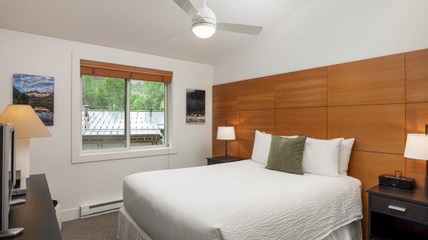 A neatly arranged bedroom with a double bed, wooden paneling, bedside tables, lamps, and a ceiling fan. Natural light enters from the window.