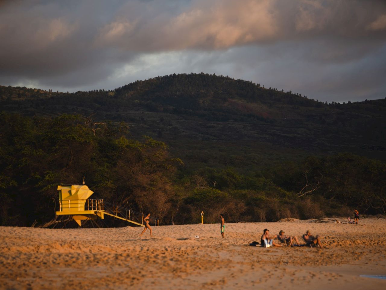 The image shows a sandy beach with a yellow lifeguard tower, people enjoying the beach, and mountains in the background.
