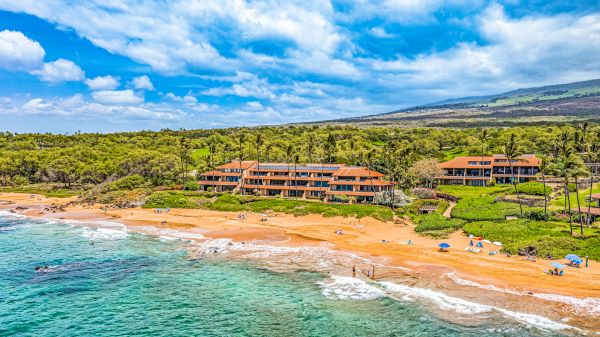 The image shows a scenic beach with clear waters, lush greenery, and beachside buildings under a blue sky with clouds.