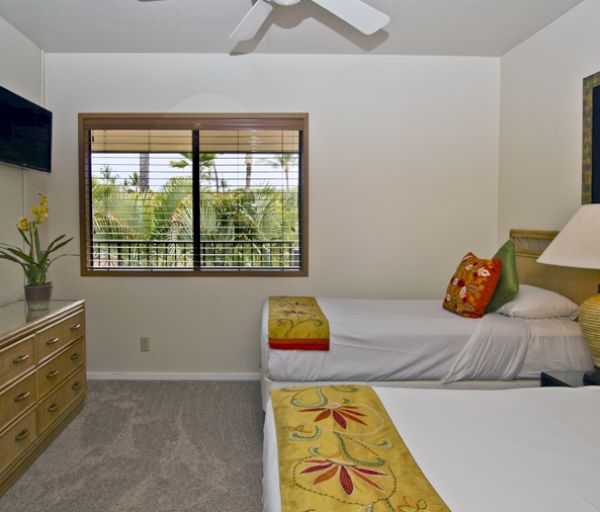 The image shows a cozy bedroom with two beds, a dresser, a TV, and a window with greenery outside, adorned with decor.
