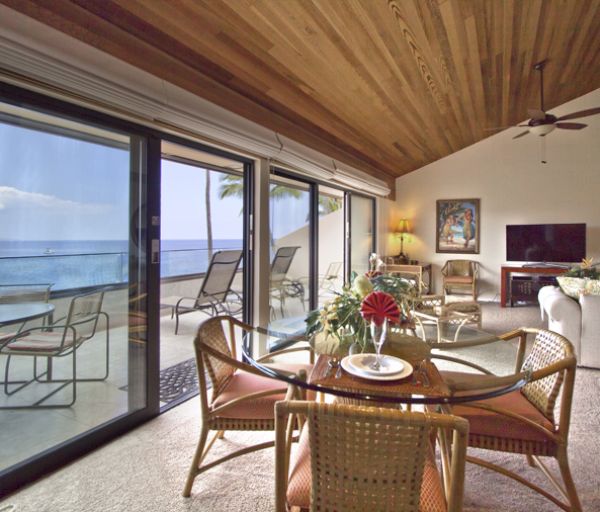 The image shows a cozy living space with large windows, a dining table, comfortable seating, and a view of the ocean.