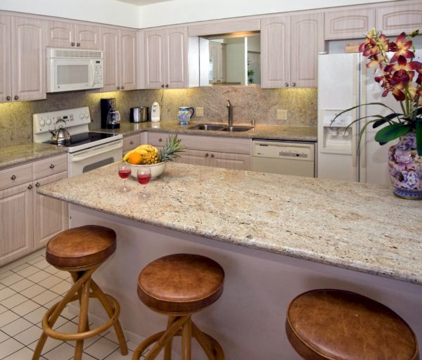 The image shows a modern kitchen with light cabinetry, a granite countertop, bar stools, and a decorative plant.