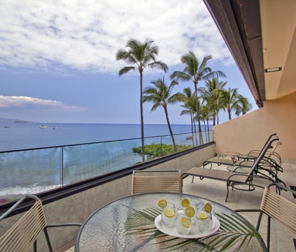 The image shows a balcony with a glass table, chairs, and a stunning ocean view with palm trees under a partly cloudy sky.