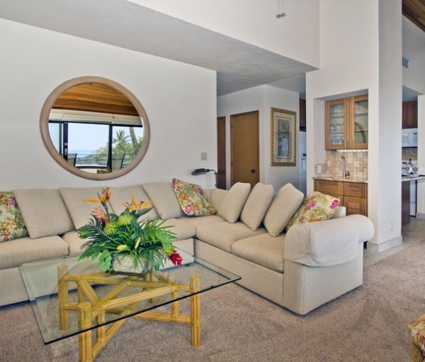 The image showcases a cozy living room with a sectional couch, coffee table, plants, and large windows allowing natural light.