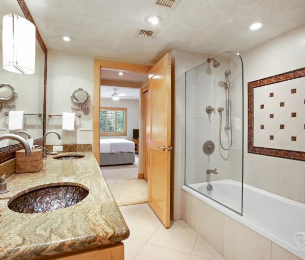 The image shows a modern bathroom with a dual sink, a glass shower, a bathtub, and a door leading to a bedroom area.