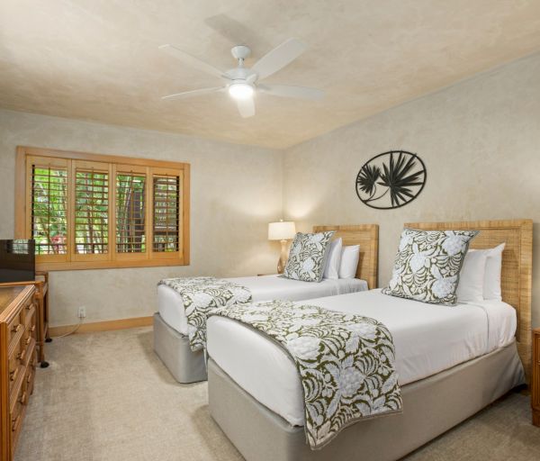 The image shows a cozy bedroom with two beds, decorative linens, a fan, wood accents, and warm lighting from lamps.