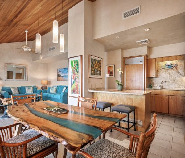 The image depicts a stylish interior with a wooden dining table, cozy seating, colorful artwork, and an inviting kitchen area.