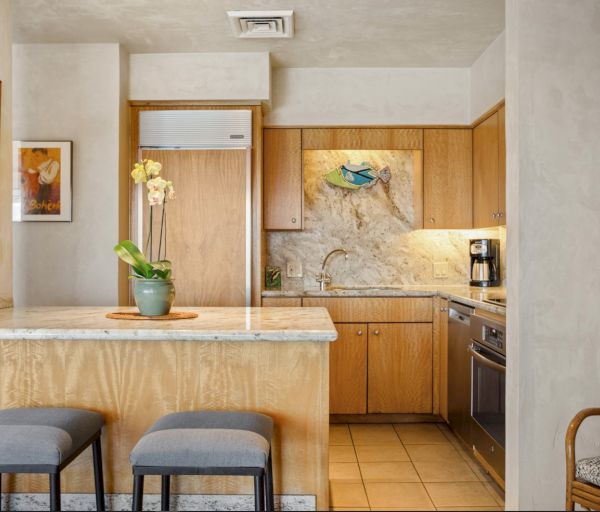 The image shows a modern kitchen with wooden cabinets, bar stools, and artwork, creating a warm and inviting atmosphere.