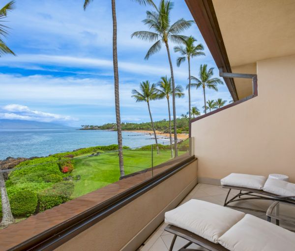 The image shows a serene coastal view from a balcony, featuring palm trees, ocean, and lounge chairs, inviting relaxation.