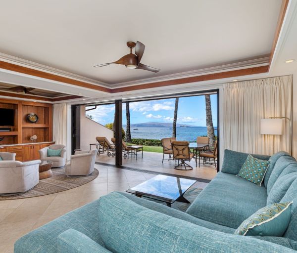 The image depicts a modern living room featuring a large teal sofa, a coffee table, and a stunning ocean view through glass doors.